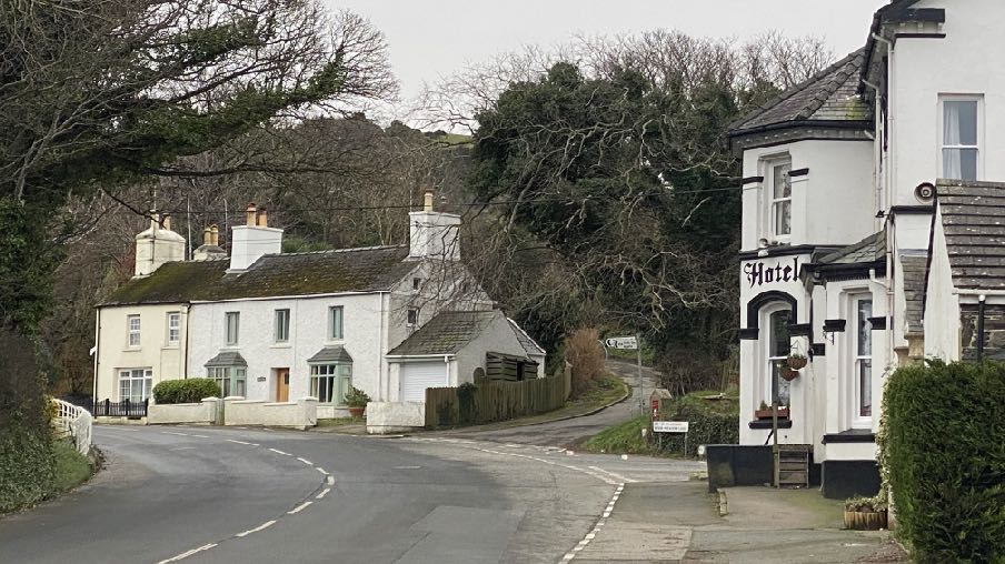 A street-level photograph showing a white two-story house with an attached garage on the left and a building with a 'Hotel' sign on the right, situated at a road junction in a rural area.
