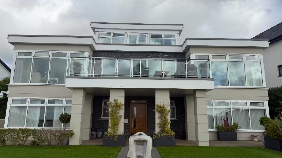 A photograph showing the front elevation of a modern, three-story detached house with a prominent first-floor balcony featuring glass balustrades and a landscaped front garden.