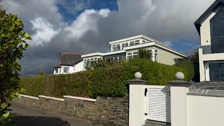 A street-level photograph showing a residential property with a stone boundary wall and tall hedge in the foreground, with modern white houses visible behind the vegetation.