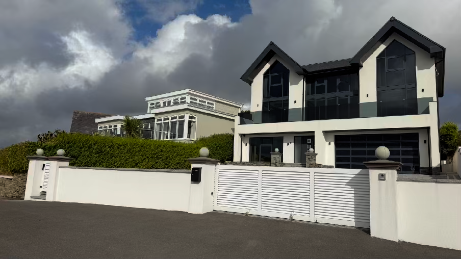 A photograph of a modern white detached house featuring a large garage door and a white boundary wall with a gate. The property is situated on a slope with another residential building visible in the background.