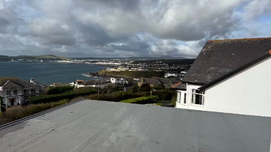 A photograph taken from a flat roof or terrace showing a white house on the right and a panoramic view of a coastal town and bay.