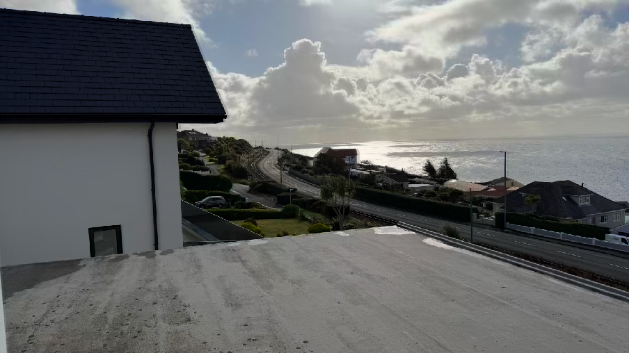 A photograph taken from a flat roof or balcony looking out towards the sea, showing the side of a white building with a slate roof on the left and a coastal road in the background.