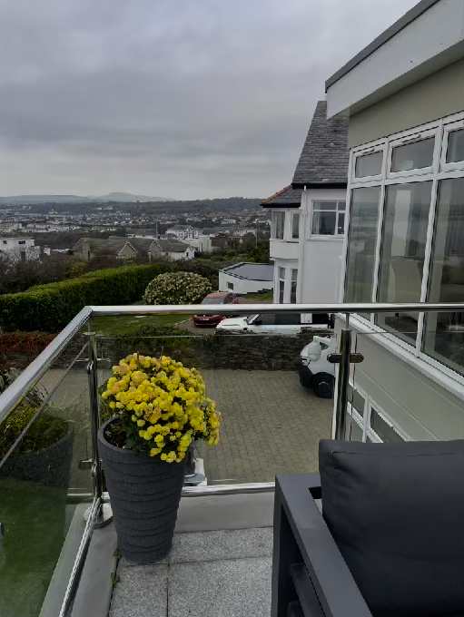 A photograph taken from a balcony showing a glass balustrade, potted yellow flowers, and a view of a residential area with parked cars below.