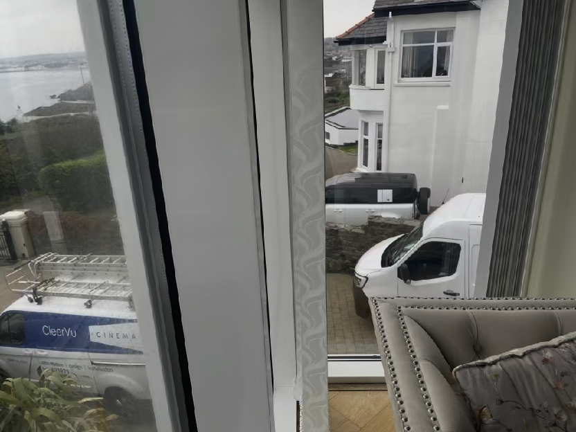 A view from inside a room looking out through a window at a neighboring white house, parked vans, and a coastal landscape.
