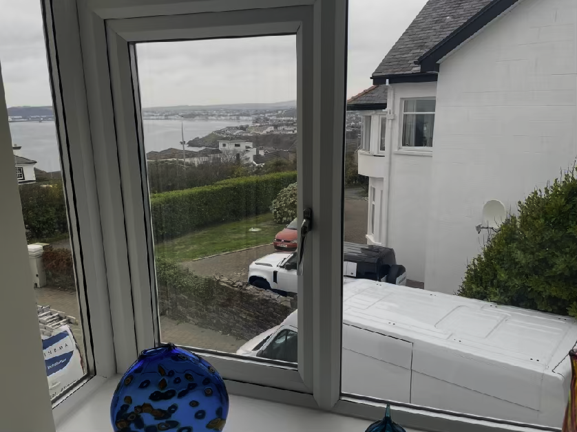 A photograph taken from inside a room looking out through a white window frame at a neighboring white house and a coastal view.