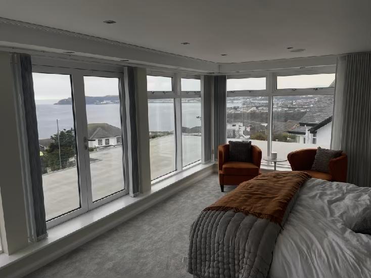 Interior photograph of a bedroom featuring large windows and glass doors overlooking a coastal view and nearby residential properties.