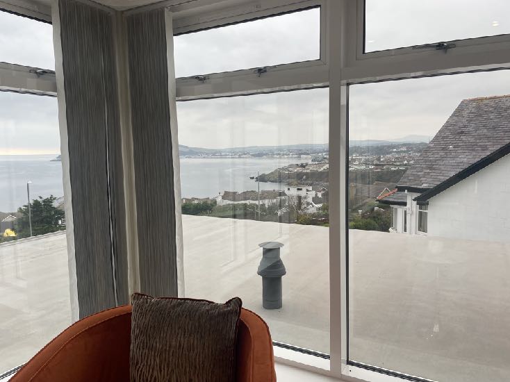 An interior photograph looking out through large windows onto a flat roof terrace and a coastal landscape.