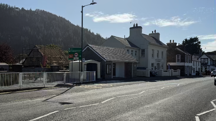 A street-level photograph showing a row of buildings including one under construction with scaffolding and a small single-story structure, set against a large wooded hill.