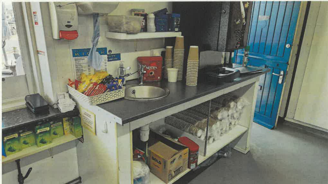Interior photograph of a kiosk service counter featuring a sink, coffee machine, stacked cups, and shelves stocked with supplies.