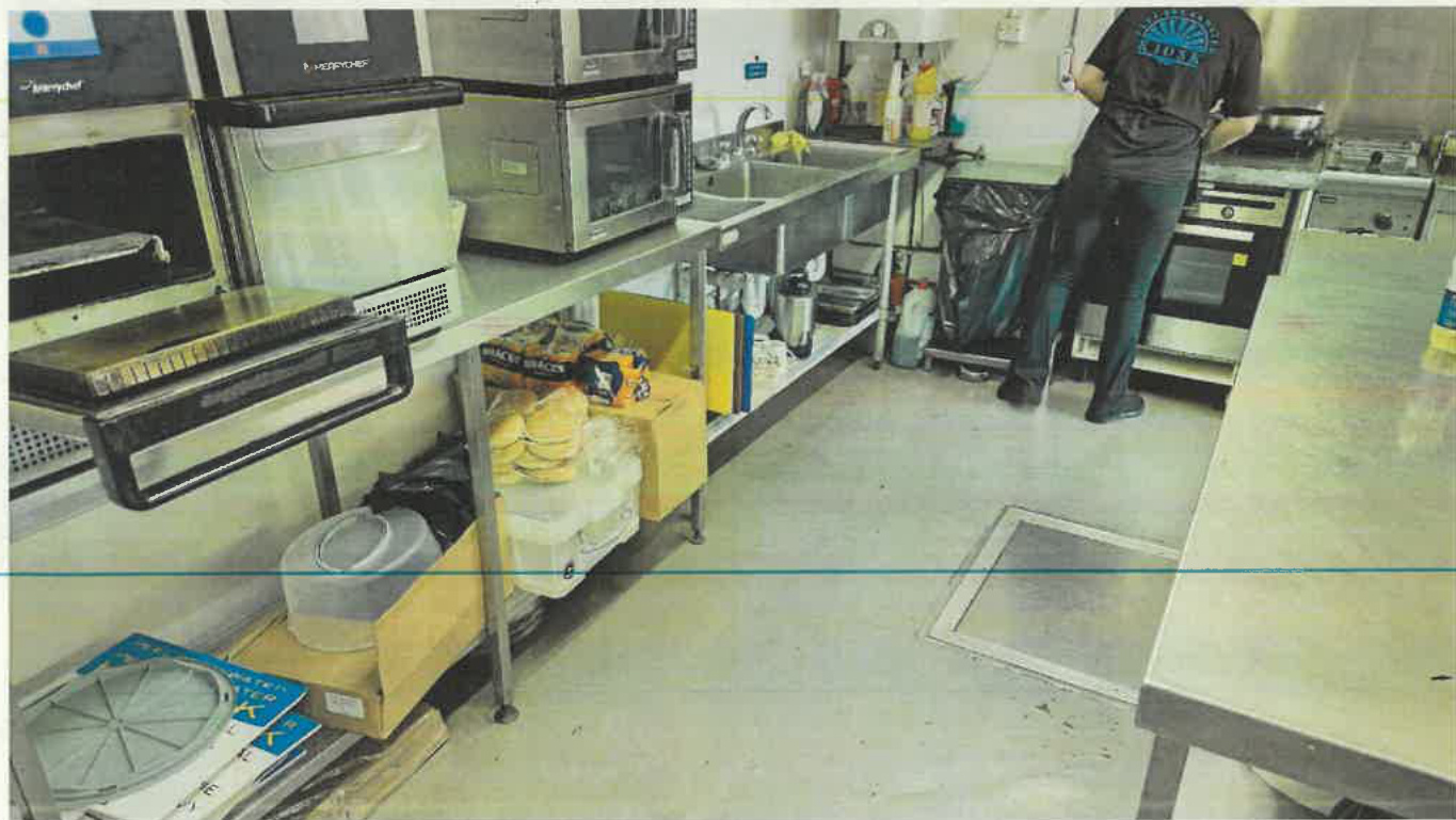 A photograph showing the interior of a commercial kitchen with stainless steel worktops, sinks, and cooking equipment.
