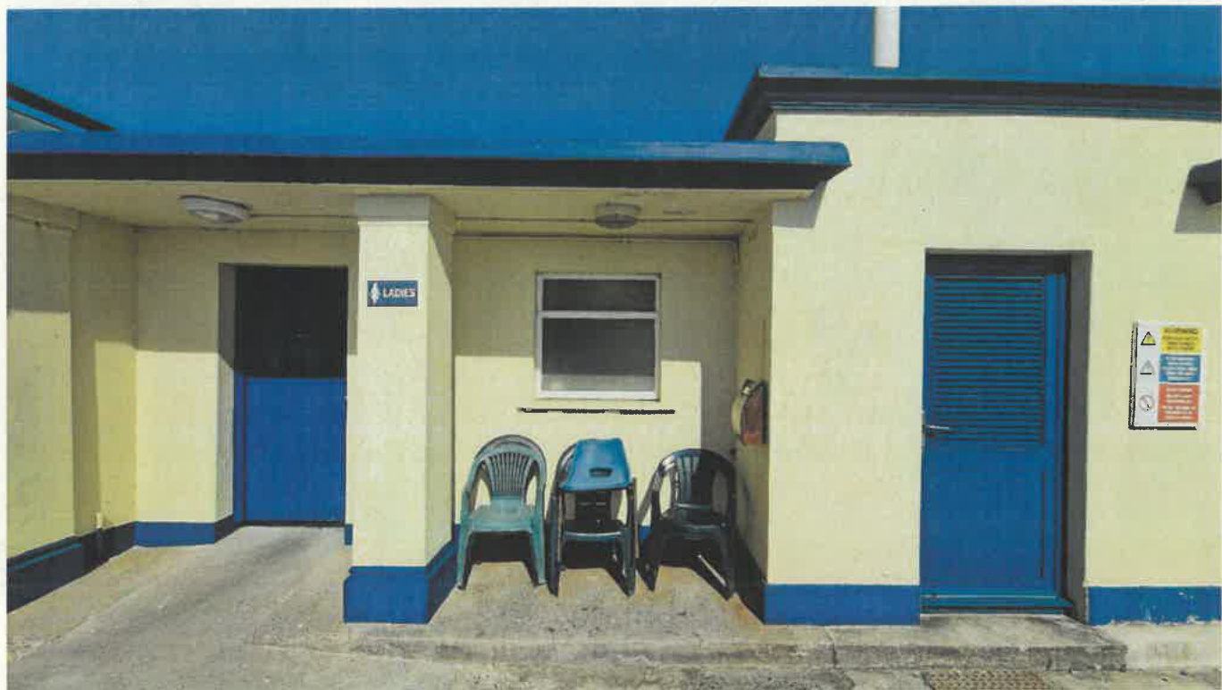 Exterior photograph of a small public toilet building with cream walls and blue trim, featuring a 'LADIES' sign and plastic chairs outside.