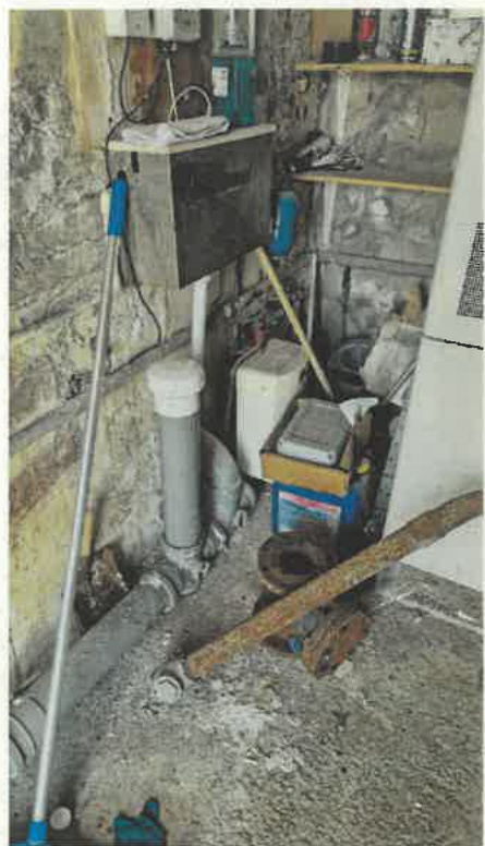 A photograph showing the interior of a utility room or storage area containing pipes, a white tank, and a metal box mounted on a stone wall.