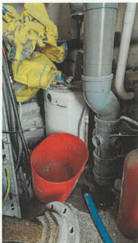 A photograph showing a utility space containing white PVC piping, a white cylindrical tank, and a red bucket.