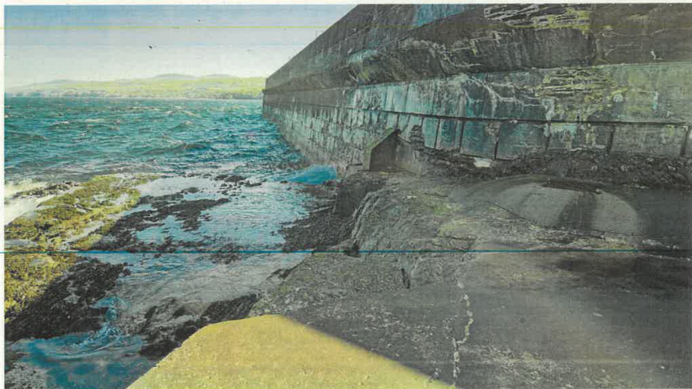 A photograph showing a coastal waterfront scene with a large stone sea wall or breakwater extending into choppy blue water and rocky shores.