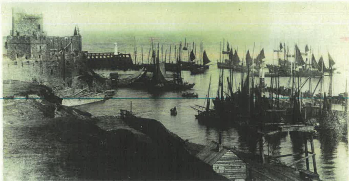 A vintage black and white photograph depicting a busy harbor scene with numerous sailing ships and a large stone castle on the left.