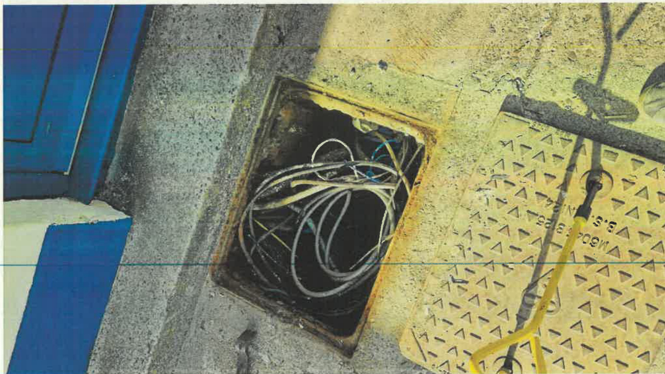 A close-up photograph showing an open utility access point or manhole containing coiled cables, situated next to a yellow metal grate and a blue painted structure.