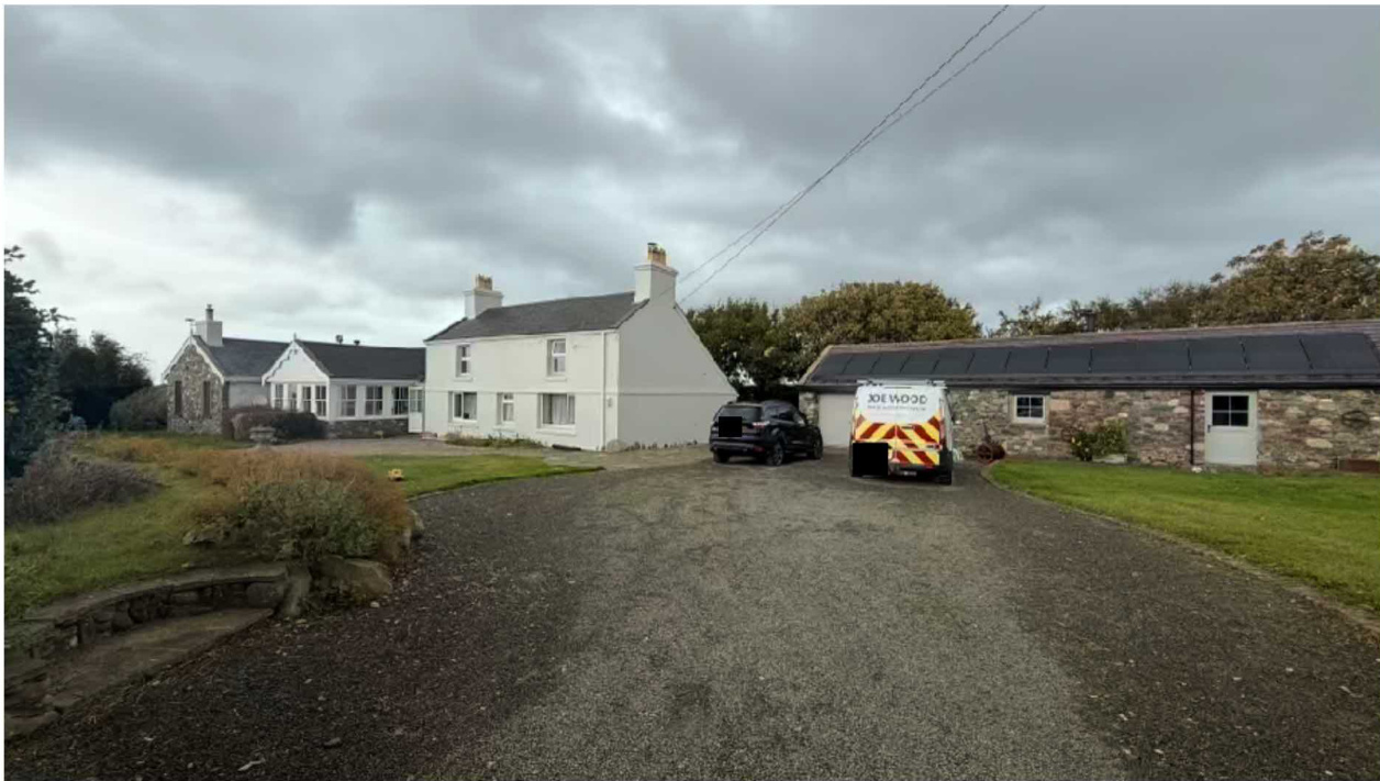 A photograph showing a rural property with a white house and a long stone building, featuring a gravel driveway with parked vehicles.