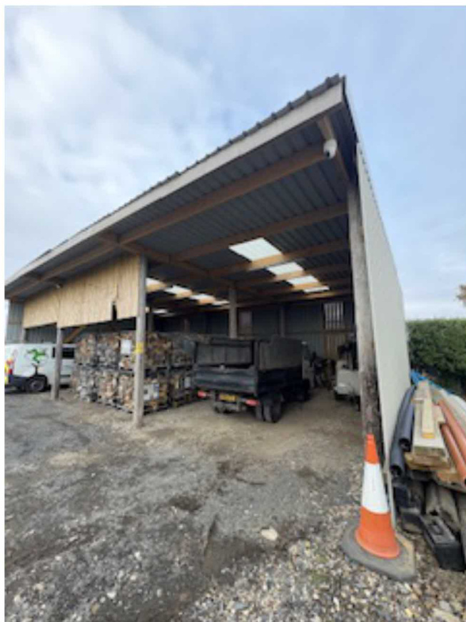 A photograph showing a large, open-sided agricultural or industrial shed with a pitched roof and skylights. The interior contains stacks of timber and a trailer, situated on a gravel surface.