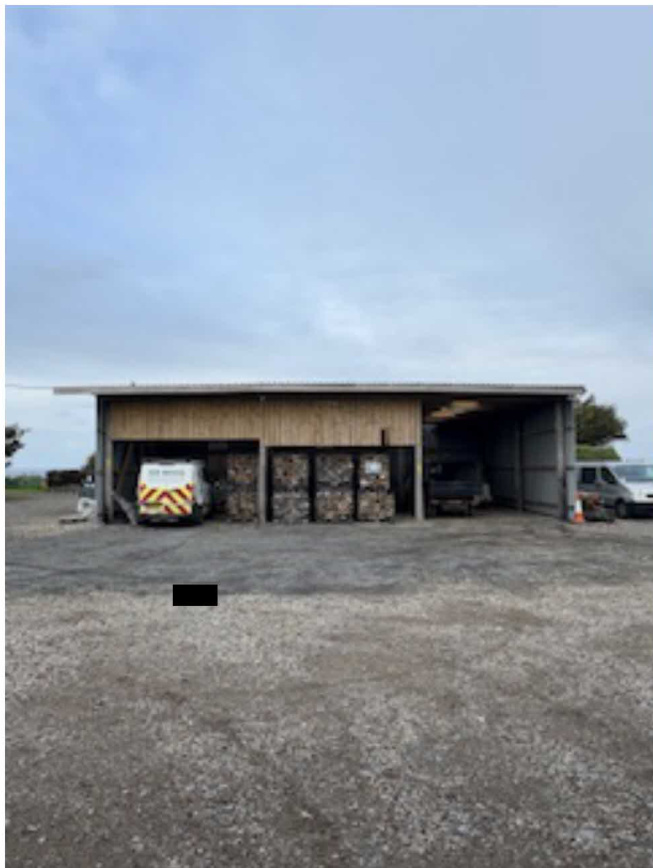 A photograph showing a large, single-story industrial building or workshop with vehicles parked on a gravel hardstanding area.