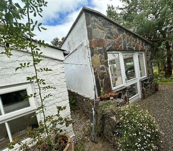A photograph showing the exterior of a property featuring a white rendered wall on the left and a stone structure with large white-framed windows on the right.