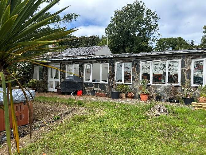A photograph showing a single-story stone building with white-framed windows and a slate roof, surrounded by a garden area with potted plants and a covered grill.