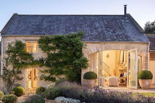 A photograph of a stone building, likely a converted barn or cottage, featuring a slate roof and large glass doors on the right side.