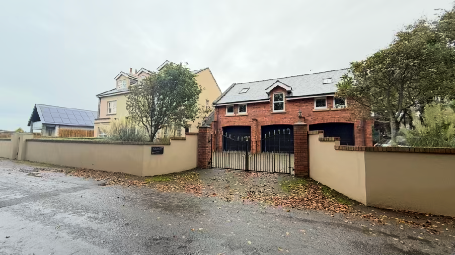 A street-level photograph showing a brick garage structure with a black metal gate, adjacent to a cream-colored house and a wall with solar panels visible in the background.