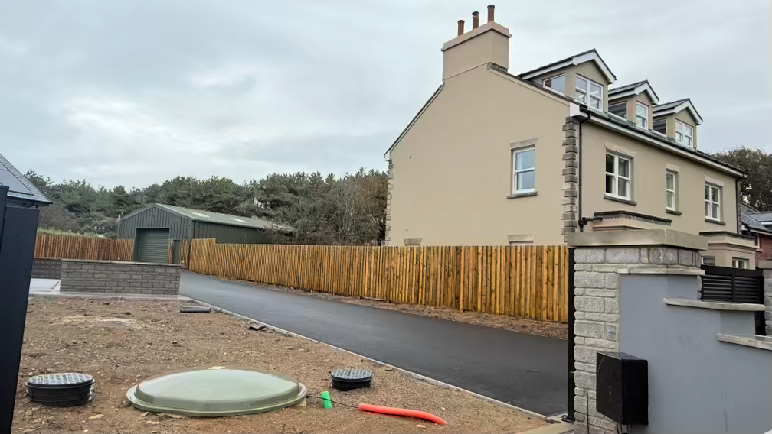 A photograph showing a residential property with a new driveway, drainage inspection chambers, and a two-story house with a wooden fence.