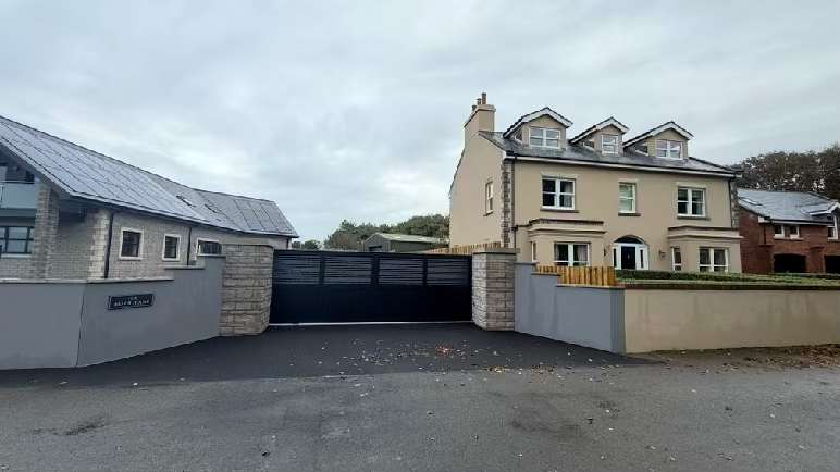 A street-level photograph showing a residential property with a modern building on the left and a detached house on the right, separated by a boundary wall and large black gate.