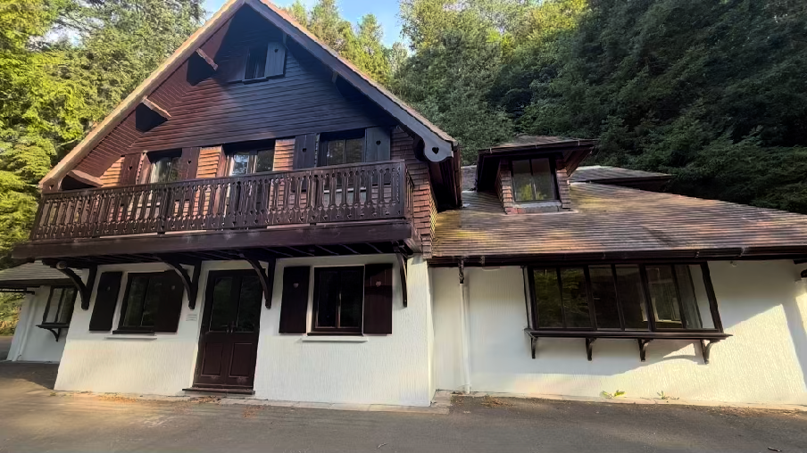 A photograph of a two-story building with dark wooden upper cladding and white lower walls, featuring a balcony and surrounded by dense trees.