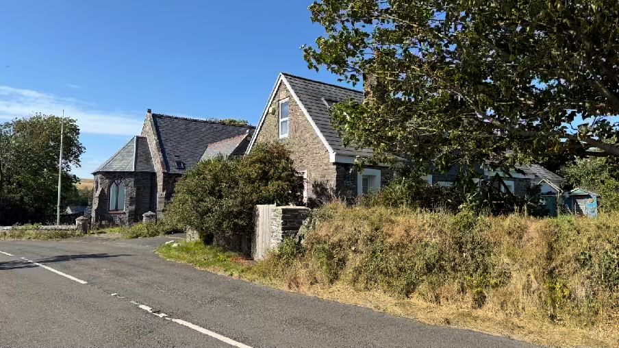 A photograph showing a stone building complex situated next to a road in a rural setting, surrounded by trees and vegetation. The image captures the exterior of the property including a stone boundary wall and overgro...