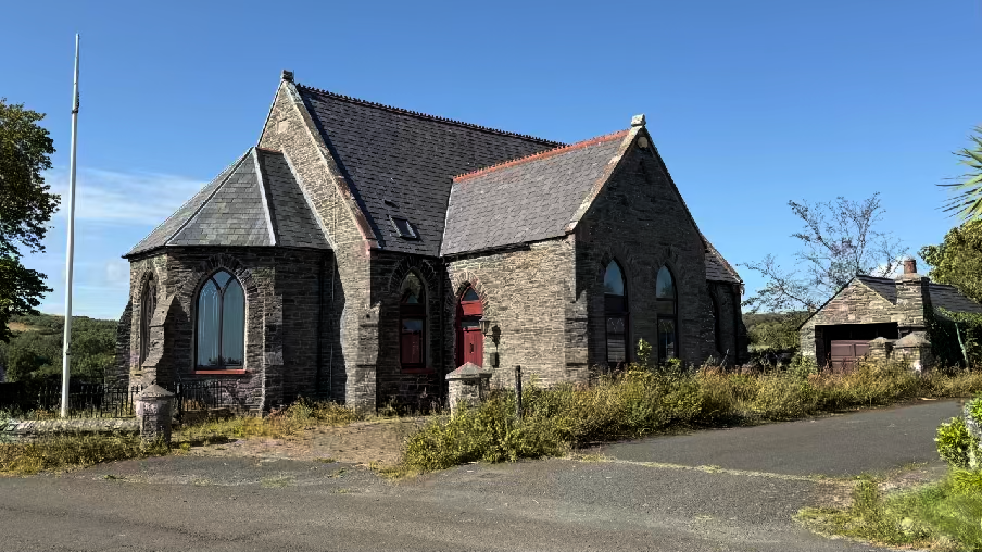 A photograph showing a stone building with a slate roof and arched windows, resembling a chapel, alongside a smaller stone outbuilding and a flagpole.