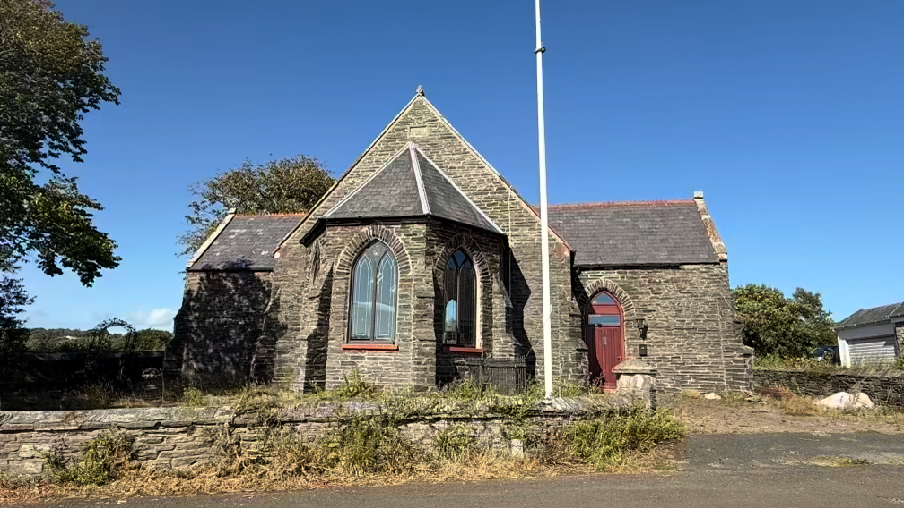 A photograph of a stone building resembling a chapel with arched windows and a red door, situated behind a low stone wall with a flagpole.