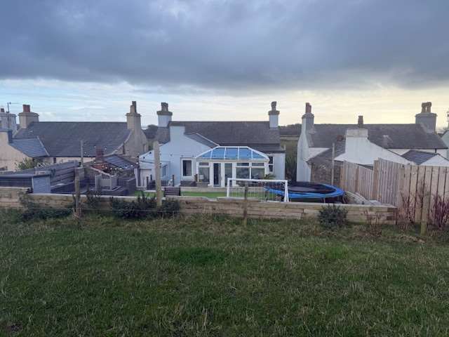 A photograph showing a row of terraced houses with a prominent glass conservatory, viewed from a grassy field in the foreground.