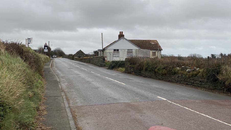 A photograph showing a rural road scene with a white, single-story dwelling on the right side, bordered by stone walls and overgrown vegetation under an overcast sky.
