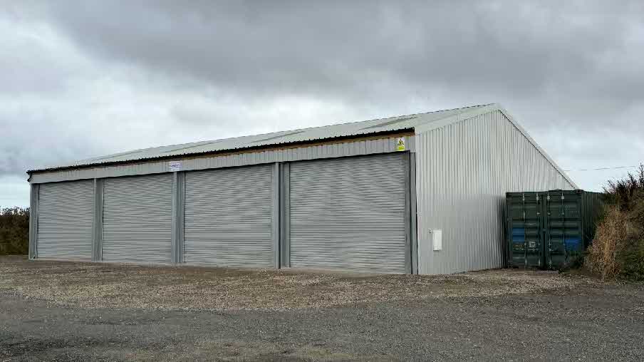A photograph showing a large, single-story industrial building with corrugated metal cladding and four roller shutter doors situated on a gravel hardstanding area.