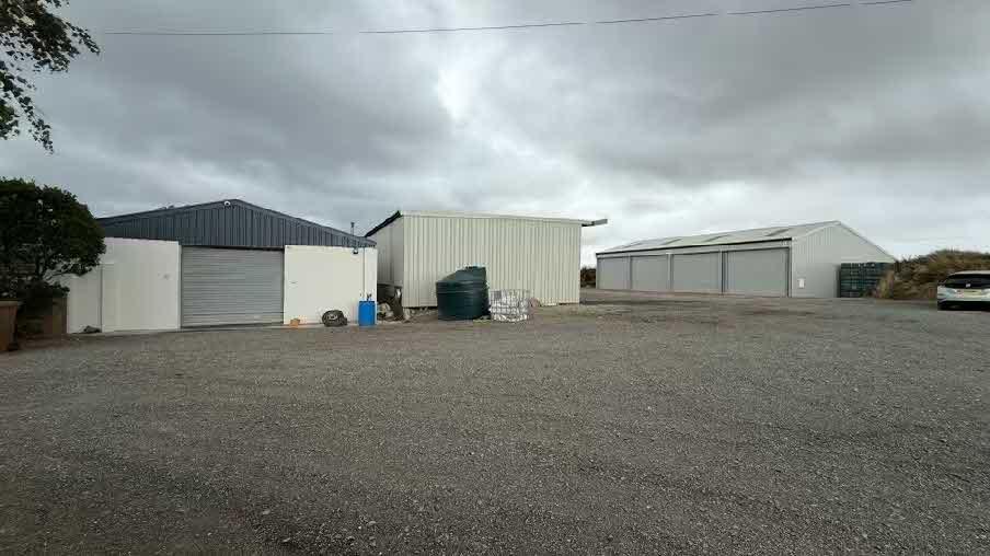 A ground-level photograph of a rural commercial site featuring three metal buildings with roller shutter doors and a large gravel hardstanding area.
