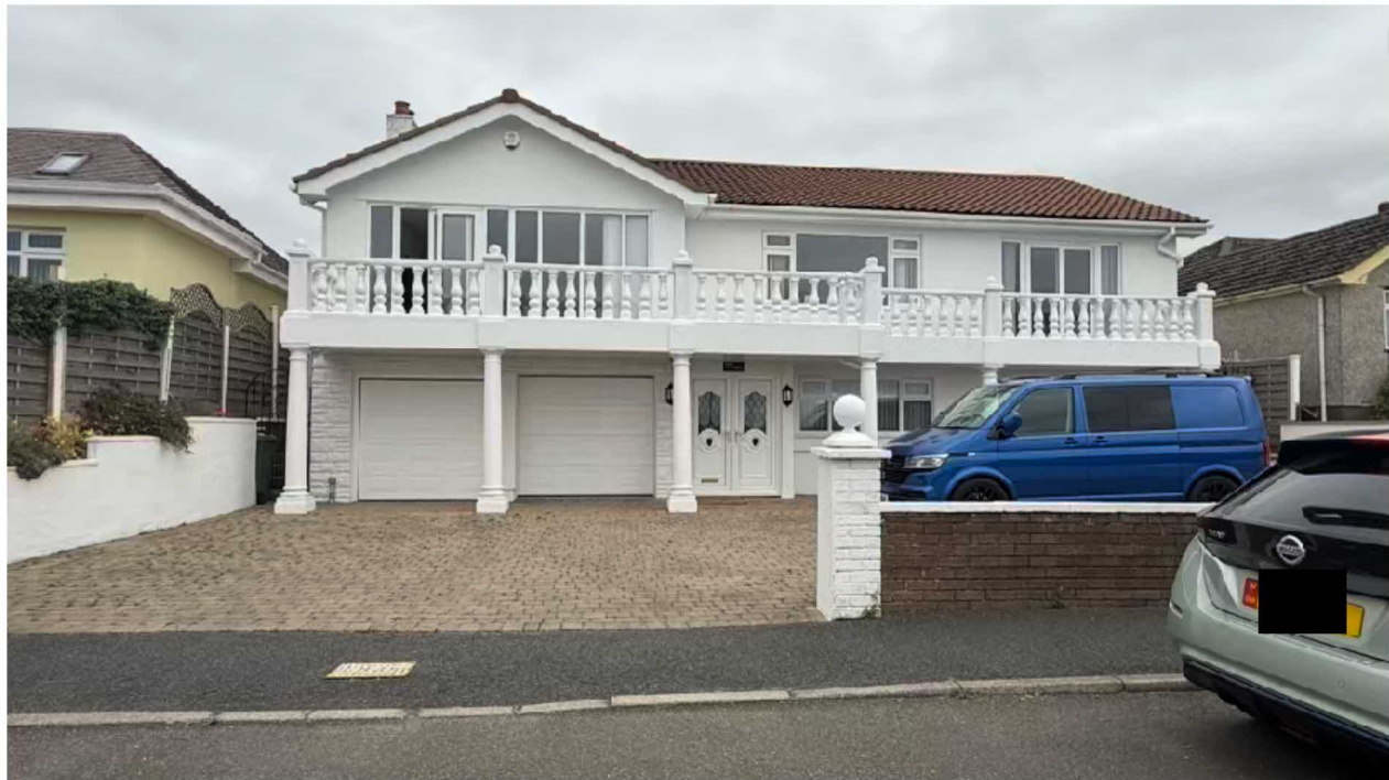 A photograph showing the front elevation of a white two-story detached house featuring a large first-floor balcony with white balustrades and a double garage on the ground floor.