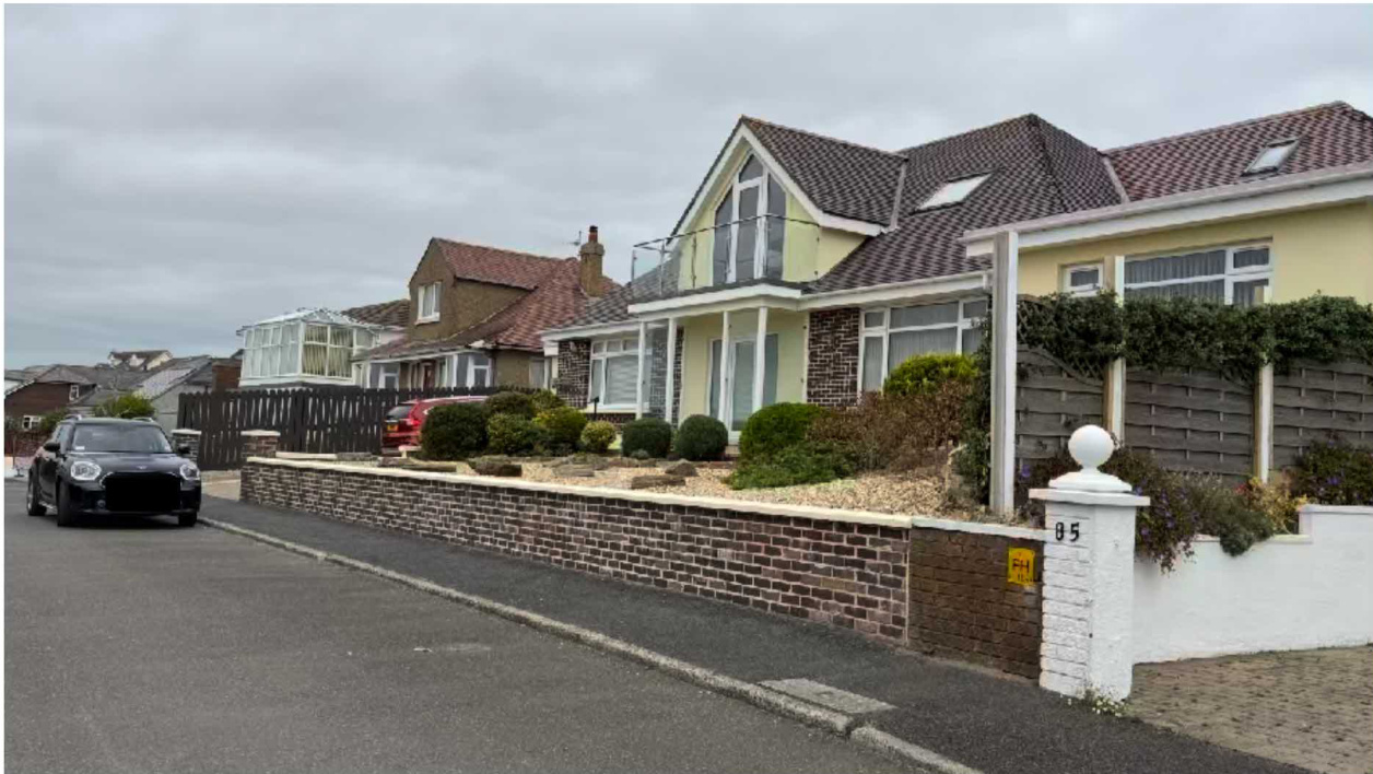 A street-level photograph showing a two-storey detached house with a glass balcony, brick boundary wall, and front garden landscaping.