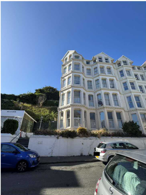 A low-angle exterior photograph of a large, white, multi-story building with Victorian-style architecture, situated on a slope with parked cars in the foreground.