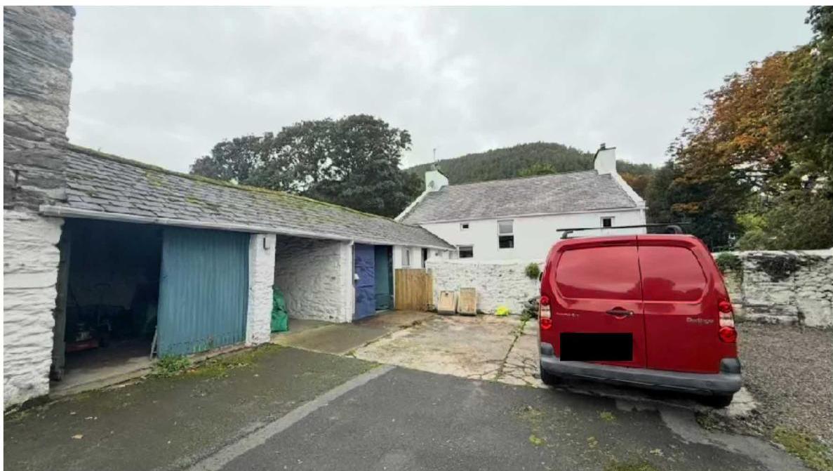 A photograph showing a rural property with a stone garage or outbuilding on the left and a white stone house in the background, with a red van parked on the driveway.