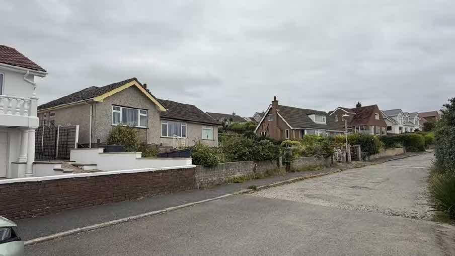 A street-level photograph showing a residential road with detached houses, featuring a bungalow with a brick boundary wall and driveway.