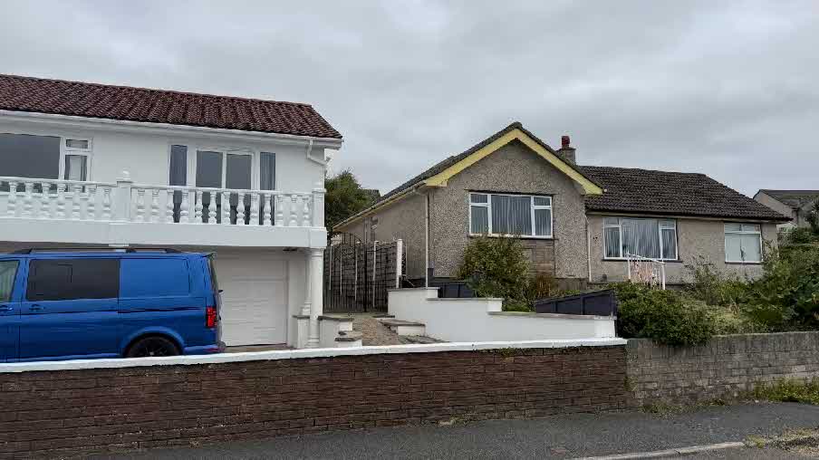 A street-level photograph showing a white two-story house with a balcony and garage next to a pebbledash bungalow, with a brick wall in the foreground.