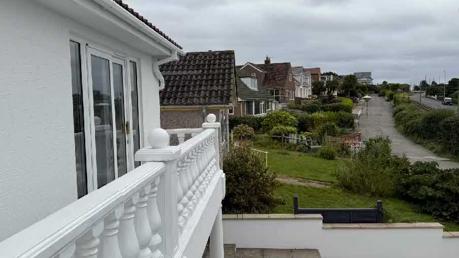 A photograph taken from a first-floor balcony showing a white balustrade and patio doors, looking out over a garden and residential street.