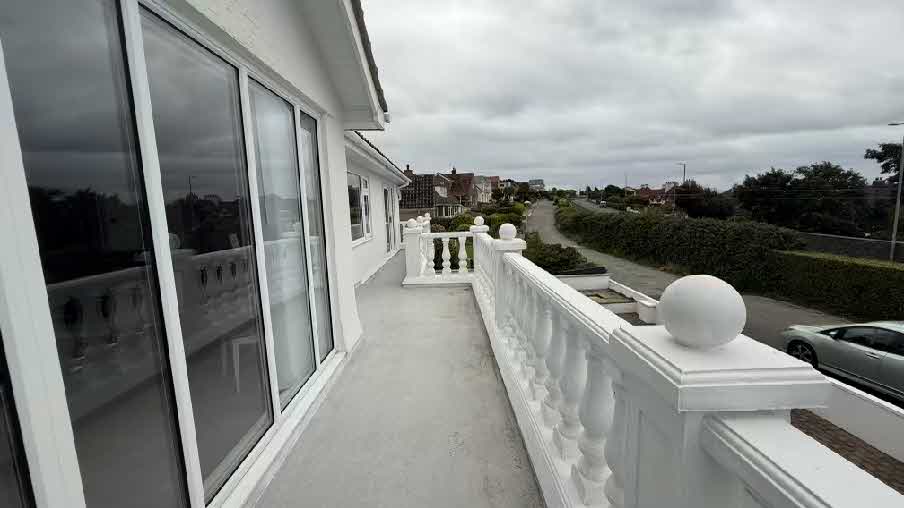 A photograph showing a first-floor balcony with white balustrades and large glass sliding doors, overlooking a residential street with hedges.