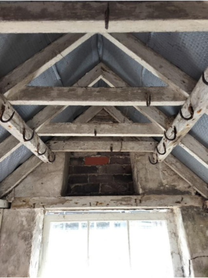 The image shows the interior roof space of a historic building, featuring exposed wooden rafters, beams, and a brick chimney breast.