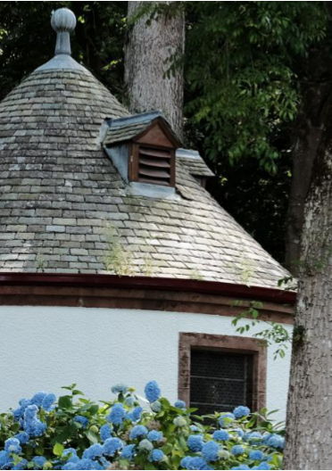A close-up photograph of a round outbuilding featuring a conical slate roof with a small dormer vent and white rendered walls.