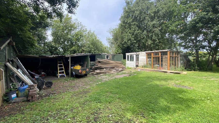 A photograph of a rural site featuring existing wooden sheds, a pile of timber, and a caravan with a wooden conservatory extension surrounded by trees.