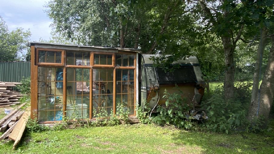 A photograph showing a wooden structure with large windows, resembling a greenhouse or sunroom, situated next to a dilapidated caravan in a grassy, wooded area.