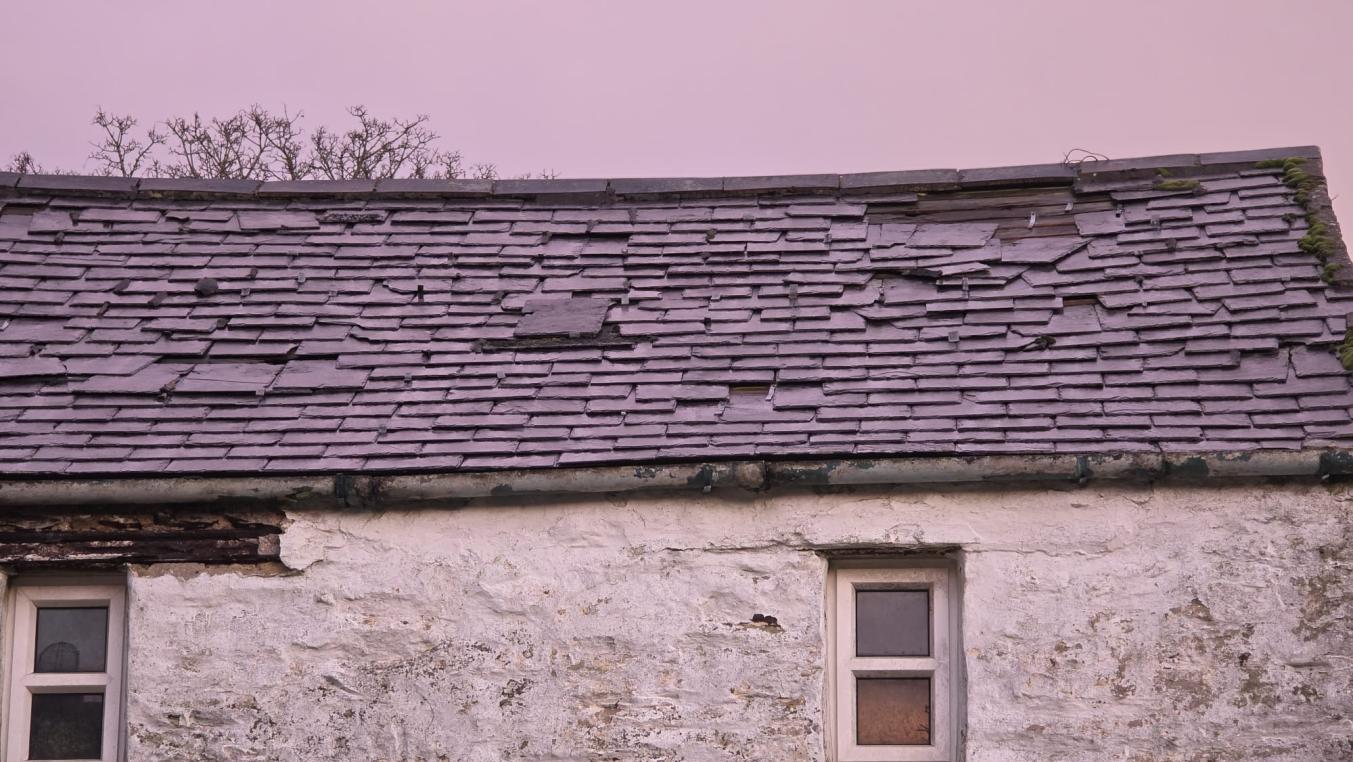 A close-up photograph showing the upper section of a weathered stone building with a slate roof and two windows.